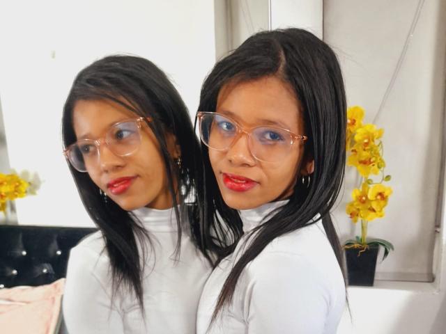 Rubyys, avec ses cheveux noirs, son rouge à lèvres rouge et ses lunettes transparentes, portant un pull blanc à col roulé, se tient devant un miroir, reflétant son visage, avec des orchidées jaunes en arrière-plan..