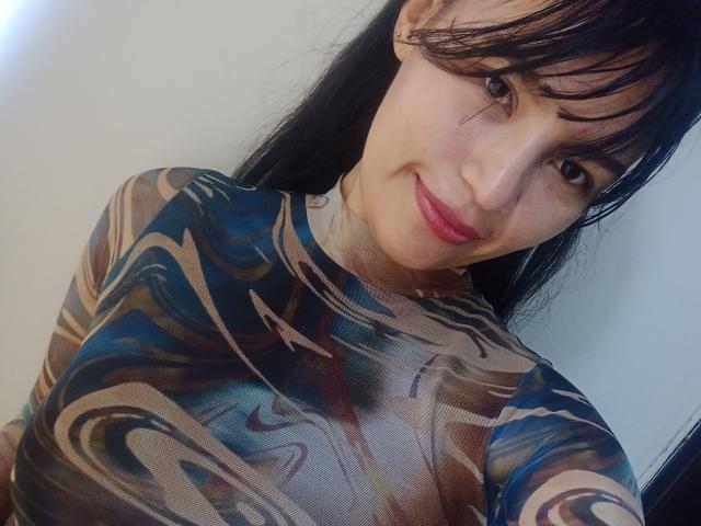 SusanKartier with black hair, bangs, and a smile, wearing a colorful, swirling-pattern, tight-fitting top, showing her boobs, has pink lipstick, and a cute, sexy look.