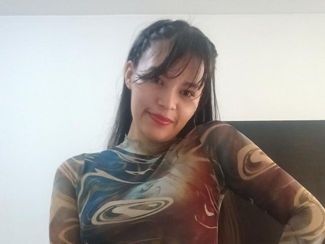 SusanKartier with black hair in braids, wearing a sheer, colorful, swirl-patterned top, smiling, showing red lipstick, and resting her hand on her ass.