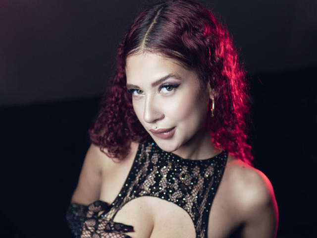 ValeryScot with curly red hair, wearing a black lace dress, covering her tits with one hand, gold hoop earrings, and a nose ring, looking at the camera with a smirk.
