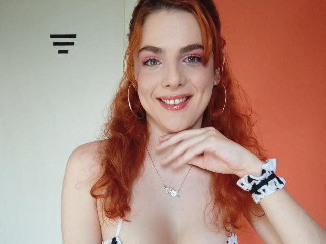 LinySex has red curly hair, pink eyeshadow, and is smiling with her hand near her chin. She's wearing a white top, hoop earrings, and a black scrunchie.