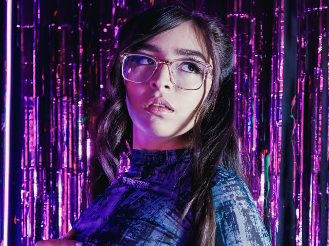 EvaMcAllister with glasses, purple-blue patterned shirt, long dark hair, purple neon lights, and a slightly open mouth, looking directly at the camera.