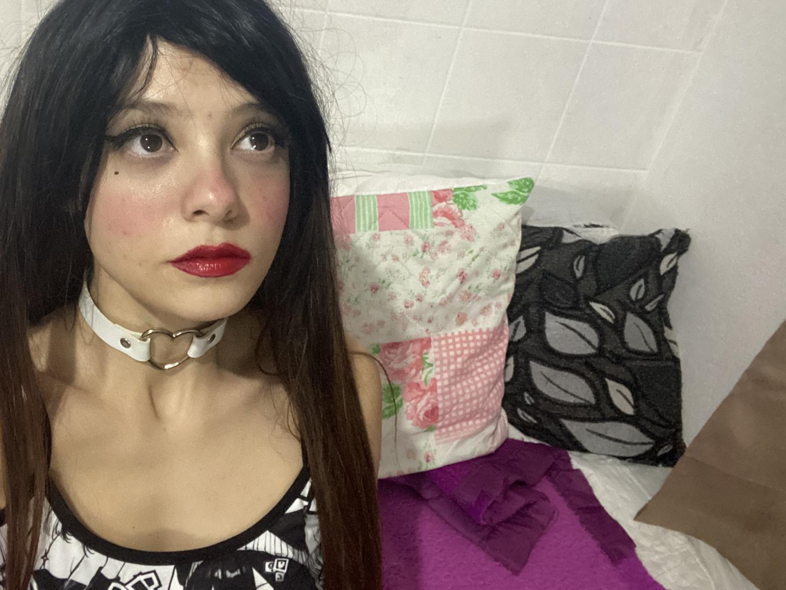 Nektafit has long brown hair, red lipstick, and a white collar with a ring. She's wearing a black and white top, sitting on a bed with pink and purple pillows.