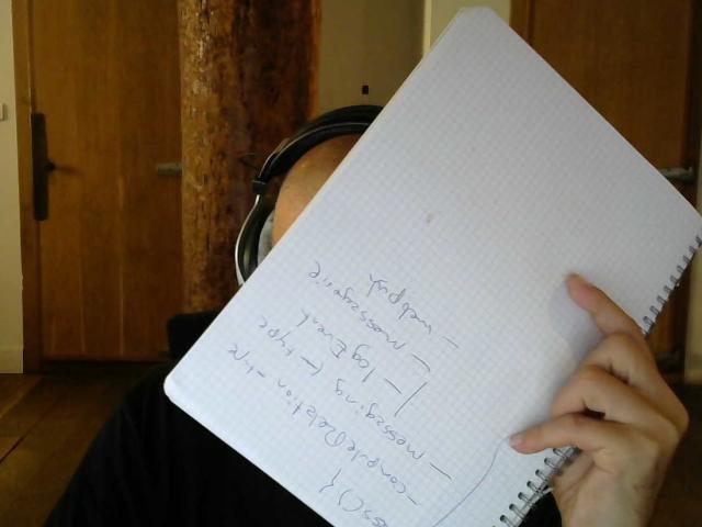Oliviermodeltest tenant un carnet devant son visage, portant des écouteurs noirs, avec seulement sa main et le carnet visibles. Le carnet recouvre ses fesses..