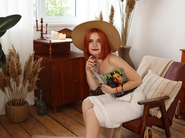 MargaritaKis, avec ses cheveux roux, portant un chapeau de paille, une robe blanche, et tenant un magazine, assise sur une chaise en bois, montrant ses seins, et souriant doucement..