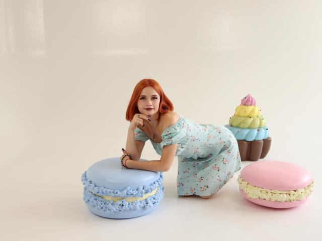 MargaritaKis with red hair, off-shoulder floral dress, kneeling on blue macaron, hand on chin, next to pink and yellow macarons, and a cupcake with frosting on white background.