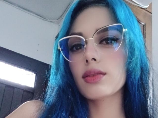 AnaisHilton with bright blue hair, wearing clear glasses, and pink lipstick, looking at the camera with a slight smile, showing her nose and lips. Her tits are not visible.
