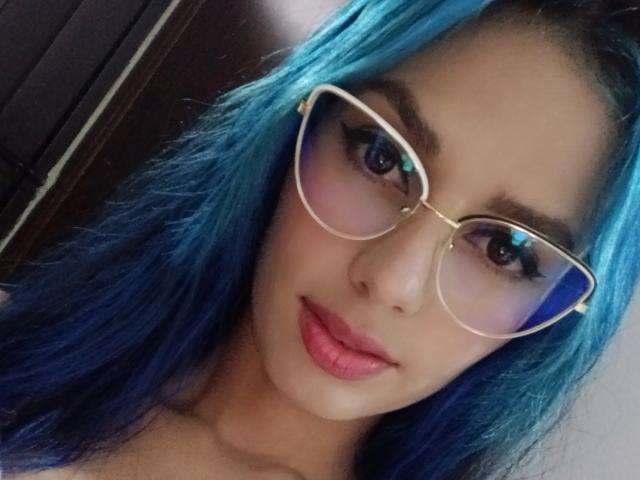 AnaisHilton with blue hair, wearing glasses, and pink lipstick, showing off her tits with a slight smile. Her eyes are blue and her skin is light.
