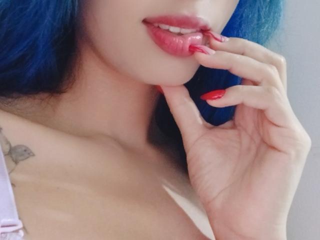 AnaisHilton with blue hair, glossy pink lips, and red nails, biting her finger, showing a small tattoo on her shoulder, with her boobs slightly visible.