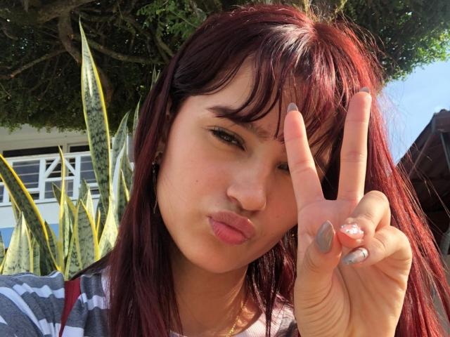 Melody Mansson, com cabelo ruivo, lábios carnudos e unhas cinzas, fazendo um sinal de paz, exibindo seu anel. Ela tem uma aparência fofa, um pouco nerd..
