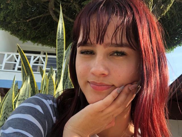 MelodyMansson with red hair, green eyes, and a cute smile, resting her hand on her cheek, wearing a grey striped shirt, standing in front of snake plants.