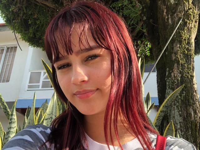 MelodyMansson has red, straight, shoulder-length hair with bangs, wearing a gray striped shirt, smiling slightly with brown eyes, and a small nose, standing in front of a tree.