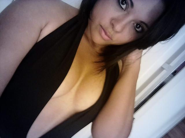 Gyxlainy with dark hair, brown eyes, and light skin, wearing a black, deep cleavage dress, looking at the camera, hand in hair, showing her tits.