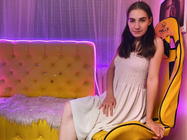 NikaStark with long brown hair, wearing a white sleeveless dress, sitting on a yellow gaming chair, legs crossed, hands on her knee, purple neon light in background.
