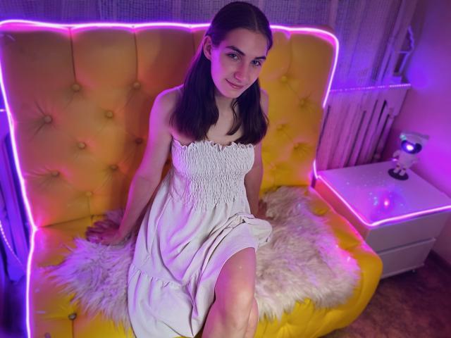 NikaStark with puffy yellow buttress sofa, neon purple trim, white dress, sitting on fluffy rug, purple and pink lighting, long brown hair, cute smile, legs crossed, wooden floor.