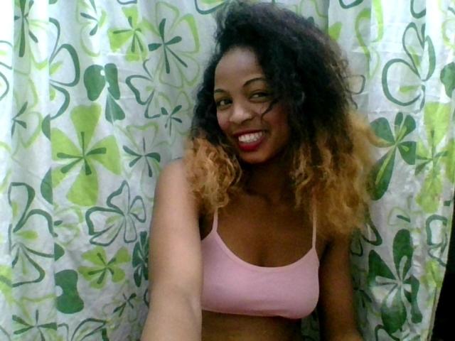 AlexaChan with curly brown hair, smiling, wearing a pink sports bra, showing her small tits, standing in front of green and white floral-patterned curtains.