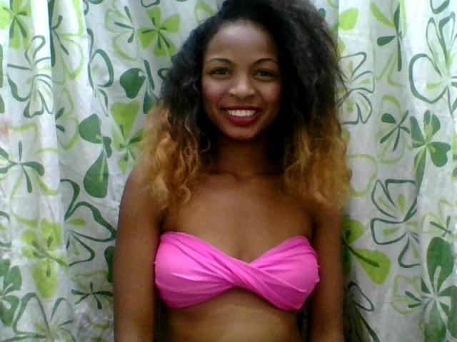 AlexaChan with curly dark brown to blonde ombre hair, smiling, wearing a pink wrap-around bikini top, showing her small tits, standing in front of green leaf-patterned curtains.