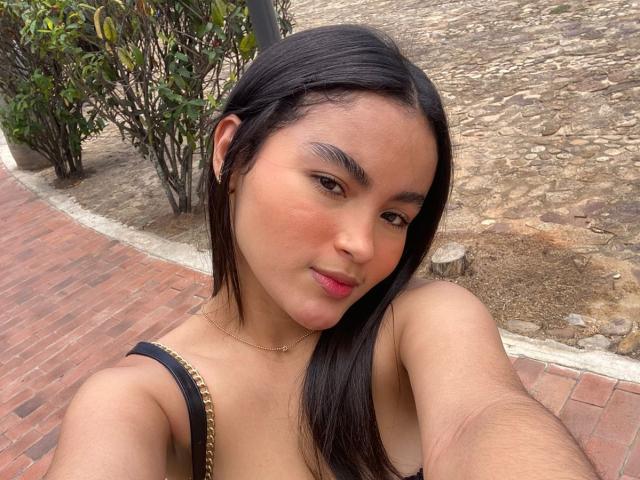 Miia Palmer, con lunghi capelli neri, pelle abbronzata, labbra rosa e un vestito nero con spalline, sorride alla fotocamera, tenendo in mano un telefono. Sullo sfondo si vedono un marciapiede in pietra e un cespuglio verde..