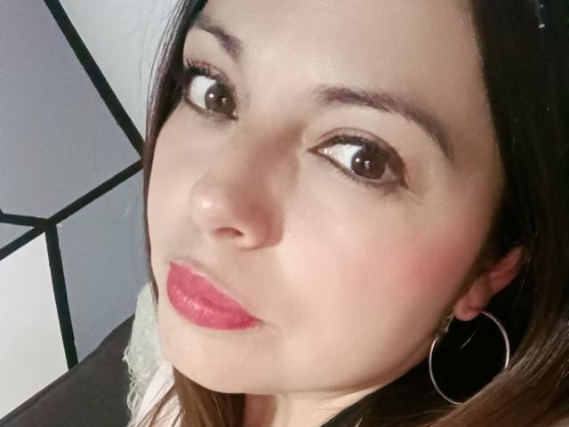 Seher's face, with brown eyes, pink lipstick, and hoop earrings, showing a slight pout. Her skin is light, and she has dark brown hair.