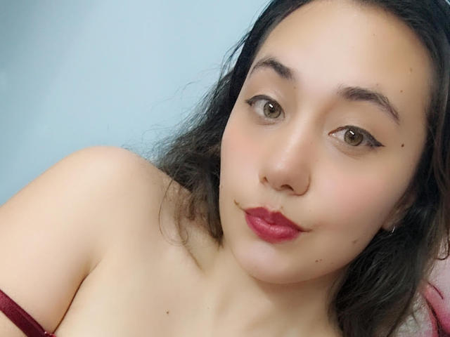 ArtemisNati has light brown skin, brown eyes, and red lipstick. She's topless with a maroon strap, black wavy hair, and a slight smile. Her tits are partially visible.