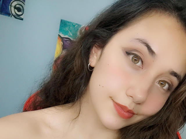 ArtemisNati with wavy brown hair, light skin, and brown eyes, wearing red lipstick and small gold earrings, smiling softly at the camera, showing a cute, sexy face.