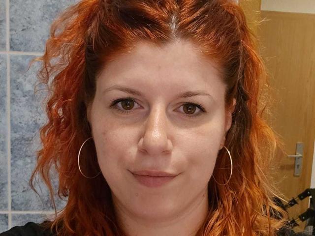 AnaysBigTits has red, curly hair, brown eyes, and is wearing thin, silver hoop earrings. Her skin is fair, and she's smiling slightly with a neutral expression.