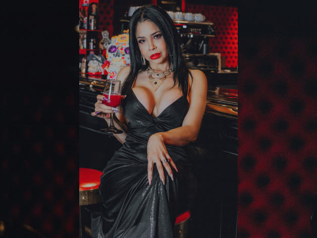 Angelina Deleon, con cabello negro, lápiz labial rojo y un vestido negro ajustado, sosteniendo una copa de vino tinto, sentada en un taburete rojo, mostrando sus grandes senos y un collar..