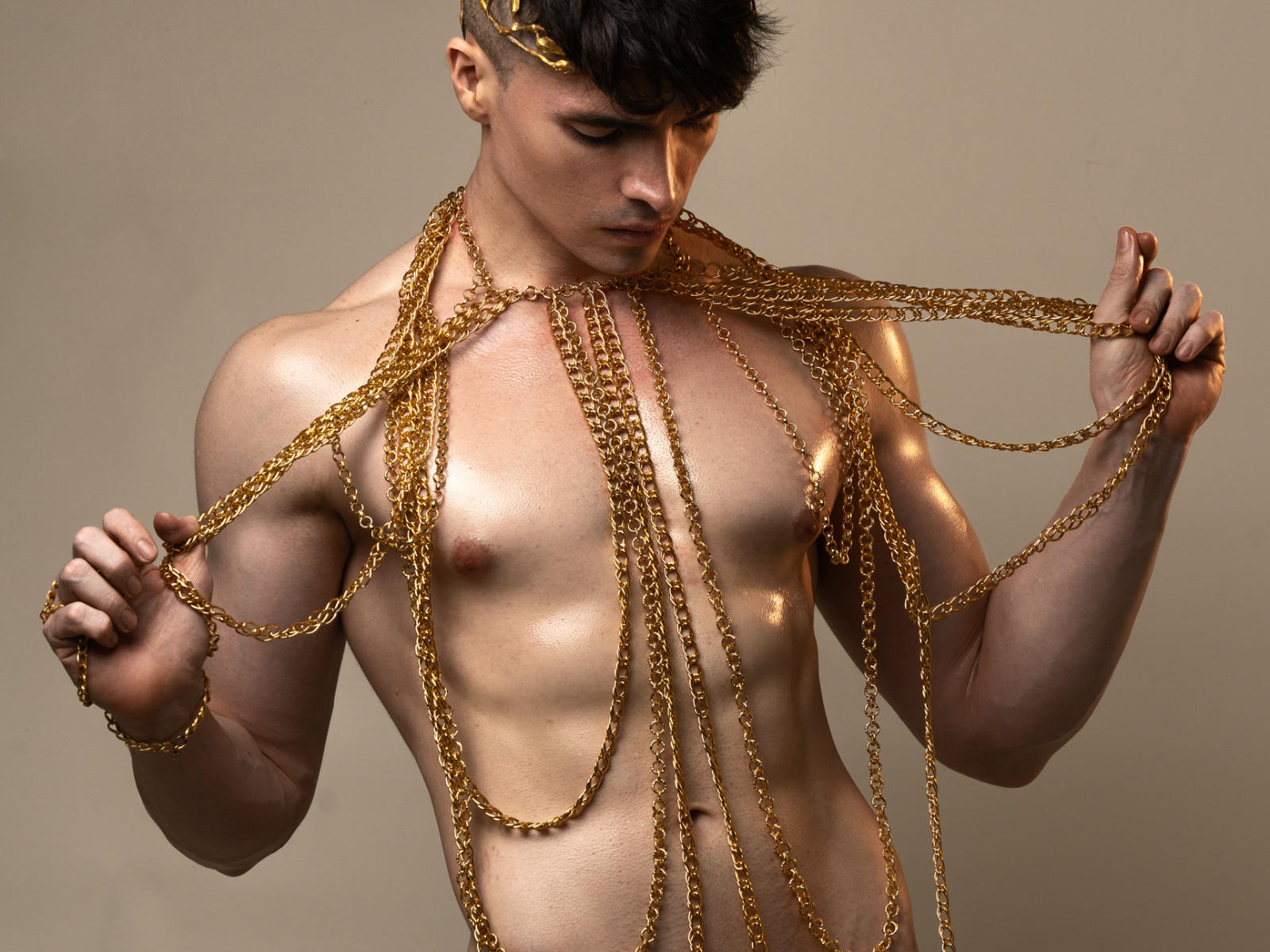 MiloLove, shirtless with a gold chain body harness, holding chains, muscular, brown hair, headpiece, looking down, beige background, shiny skin, toned abs, attractive, male, sexy.