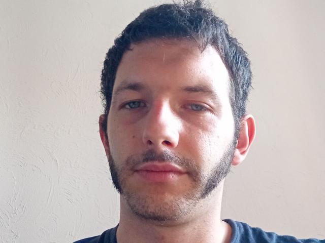 El rostro de Aure69, con cabello negro rizado y corto, barba clara y un ligero bigote, con una expresión neutra y piel clara, vistiendo una camisa negra..