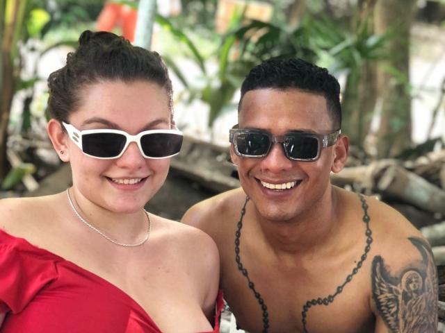 EvaAndMarcelo, wearing white sunglasses and red dress, smiling with Marcelo, shirtless, black chain necklace, tattoos, brown skin, black sunglasses, both outdoors, Eva has white frame glasses, Marcelo has short hair.