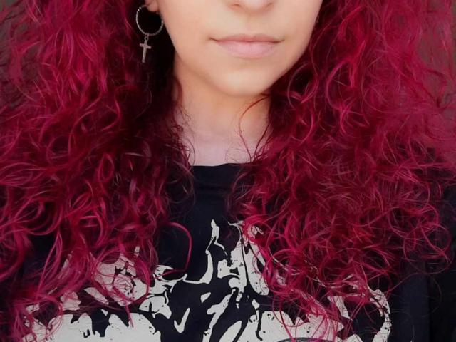 Agaa with curly red hair, wearing a black shirt with white patterns, and a black cross earring, showing her pale skin and subtle smile.