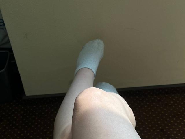 Le gambe chiare di Valissia, con calze bianche, una gamba sollevata contro un muro beige, che mostra il suo sedere scoperto e le cosce superiori in una stanza con tappeto..