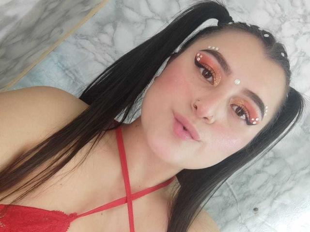 WetHairyPussyX with black pigtails, red eye makeup, nose ring, and red lace bra, showing pink pout, white forehead dots, and red strappy top. Marble wall background.