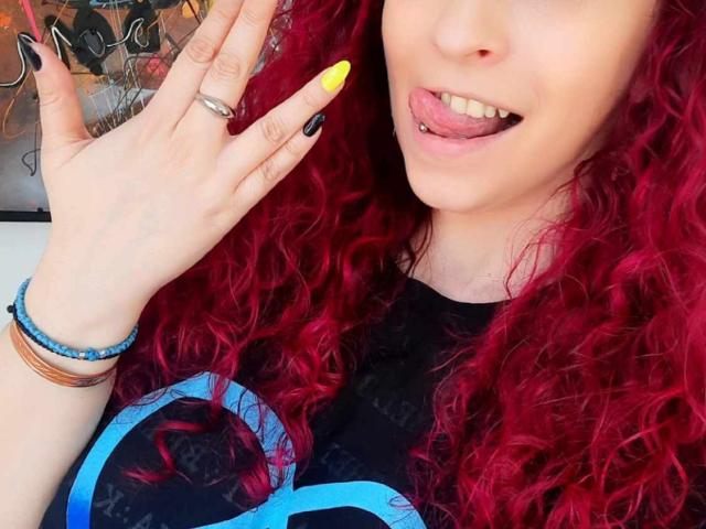 Agaa with red curly hair, yellow nail, black nail, tongue out, silver ring, and bracelets, showing off hand. She's got a cute face and a sexy look.