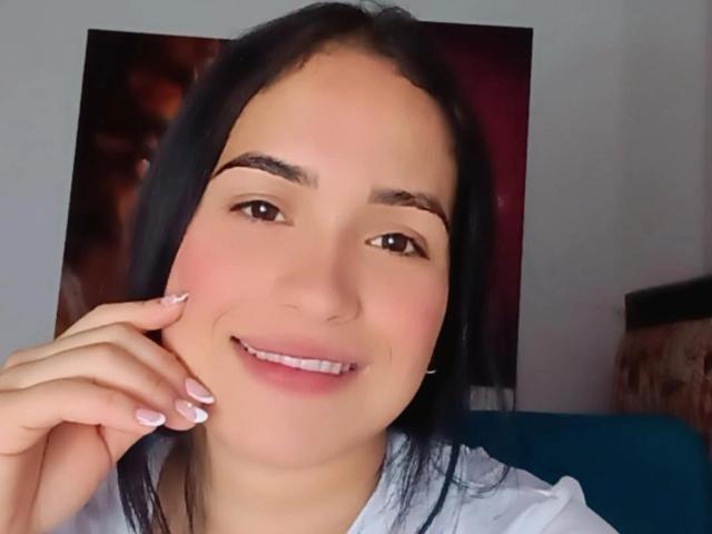MartinaDupont with black hair, brown eyes, and pink lips, smiling with hand touching cheek, wearing white shirt, showing pink manicured nails, and having cute white teeth.