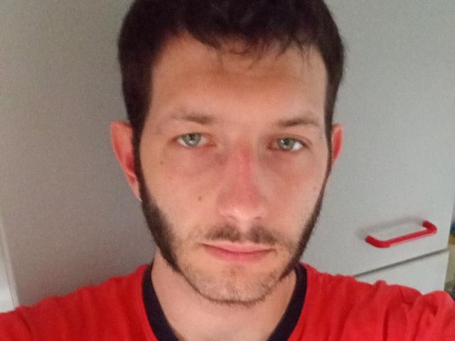 Aure69, con capelli neri corti, occhi verdi, barba leggera e camicia rossa, guarda dritto verso la fotocamera con un'espressione leggermente neutra. Il suo naso è leggermente rosso..