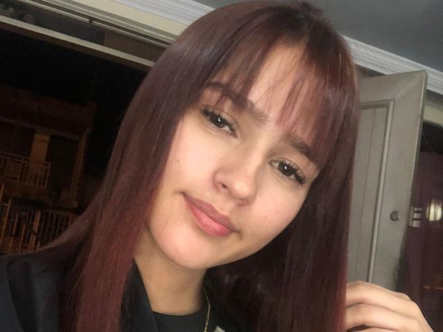 MelodyMansson has straight, red-brown hair with bangs, light skin, and a slight smile. Her brown eyes are half-open, and she's wearing a black top.