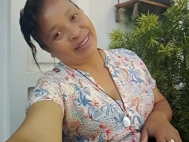 Rahel69 with a cute smile, wearing a patterned pink and blue shirt, has her dark hair in a ponytail, and a necklace with a white button. Her tits are small.