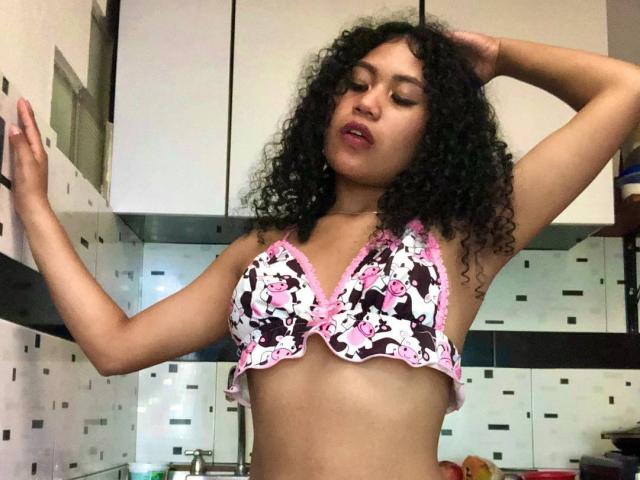 SophieSonyas with curly black hair, wearing a floral bra with pink trim, standing in a kitchen, arms raised, eyes closed, looking slightly seductive, showing her small tits.