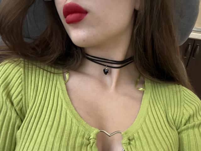 ElMarina with red lipstick, green ribbed top, black choker with black heart, and brown hair, showing cleavage and collarbone. Boobs partially visible.