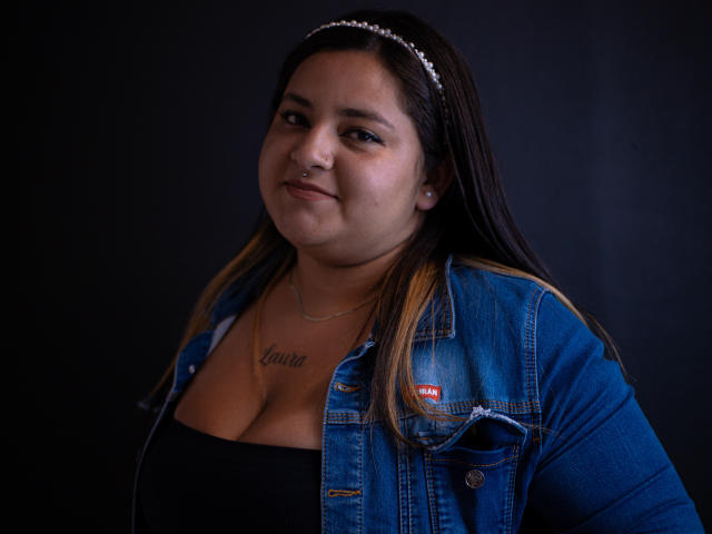 DakotaDupont, a chubby woman with long brown hair, wearing a denim jacket and black top, has a nose ring and tattoo under her chest, smiling softly.