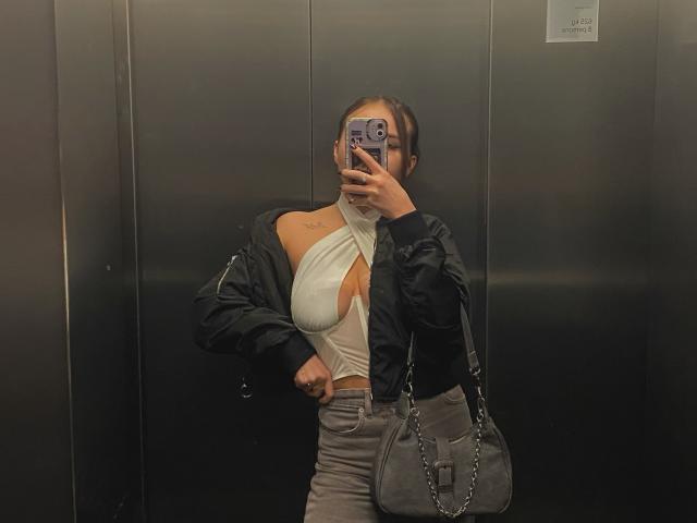 BrendaMoonx with big tits, wearing a white halter top, black jacket, gray pants, and gray bag, taking a selfie in an elevator, phone covering her face.