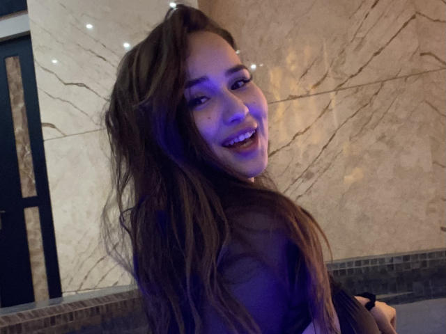 BrendaMoonx with long brown hair, smiling at the camera, blue light on her face, wearing a dark top, sitting in a tiled area, looking over her shoulder.