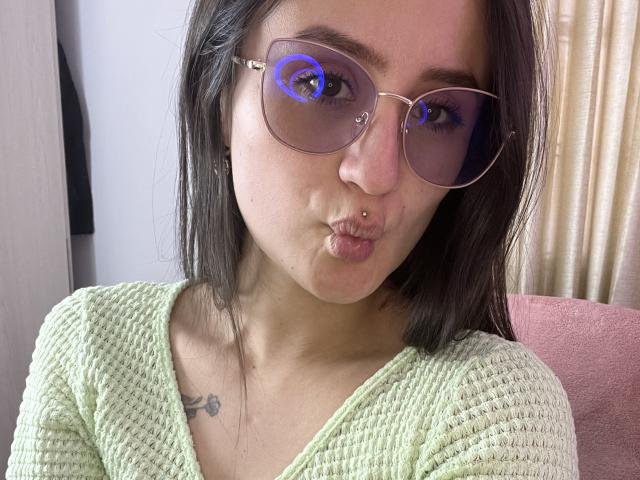NinaBryons with pouty lips, wearing green knit top, and pink-tinted glasses with blue ring on lens, has nose ring and tattoo on left chest. Dark hair, fair skin.