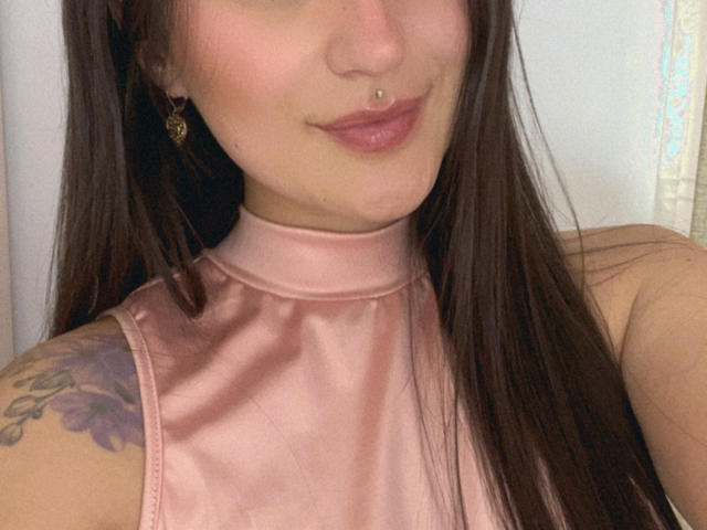 NinaBryons with long brown hair, pink satin top, nose ring, and a tattoo on her left shoulder, showing a subtle smile. Her pink lips are slightly parted.