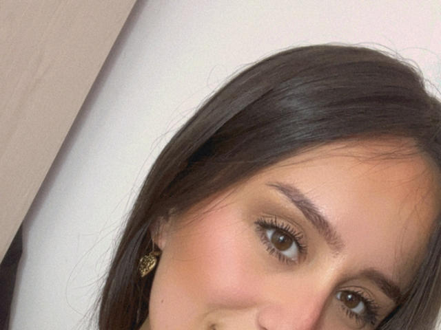 NinaBryons has brown eyes, dark straight hair, and wears gold earrings with a butterfly design. Her face is slightly tilted, showing a subtle smile.