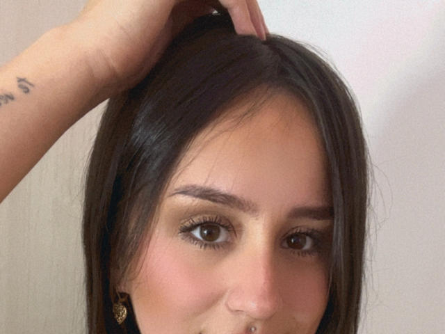 NinaBryons has brown eyes, long brown hair, and a nose ring. Someone's hand is gently touching her head. She's wearing gold earrings. Her face is slightly flushed.