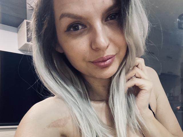 DouceAmelia has light brown skin, medium-length blonde and gray hair, and pink lipstick. She's smiling with a hand near her face, showing a small scar on her shoulder.