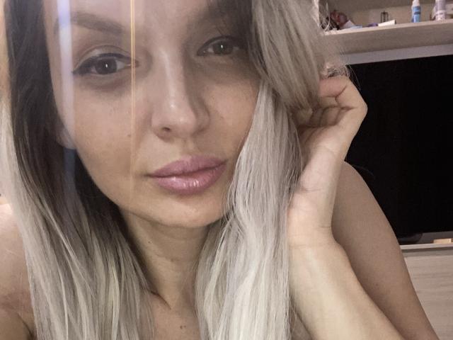 DouceAmelia with blonde hair, pink lips, and a ring on her right hand, looking at the camera with a slight smile, her hand near her head.