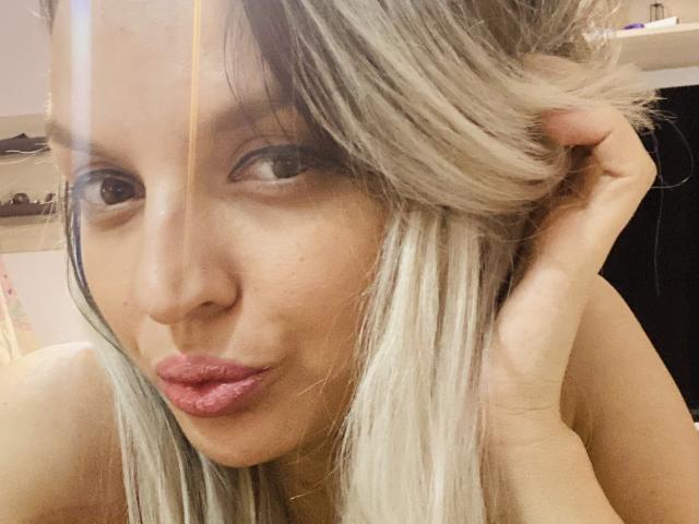 DouceAmelia with pouty pink lips, blonde hair, and brown eyes, touching her hair with one hand, looking at the camera with a slight smile.